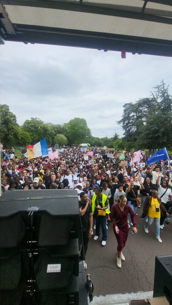 Vue d'une grande foule lors d'un événement, avec des drapeaux et des pancartes, devant du matériel de sono.