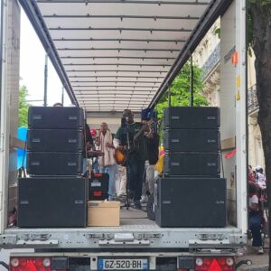 Concert en plein air dans un camion avec des musiciens et du matériel de sono visible.