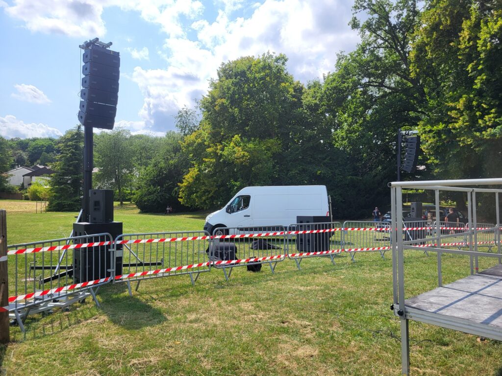 Matériel de sono installé dans un parc pour un événement, avec un camion et des barrières de sécurité en arrière-plan.