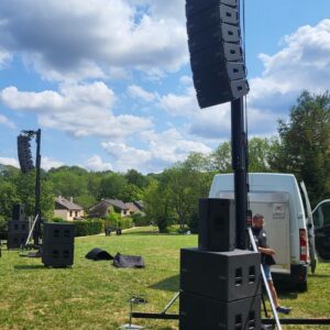 Installation de matériel de sonorisation sur un terrain verdoyant, avec des hauts-parleurs alignés et un véhicule utilitaire à proximité.