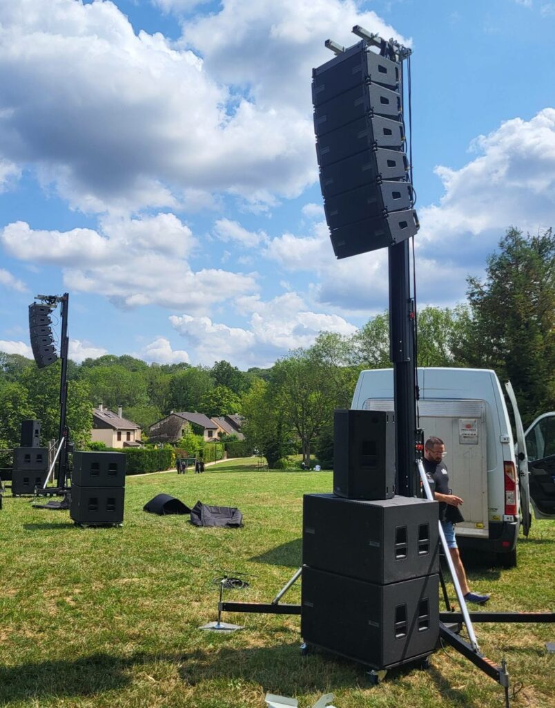 Installation de matériel de sonorisation sur un terrain verdoyant, avec des hauts-parleurs alignés et un véhicule utilitaire à proximité.