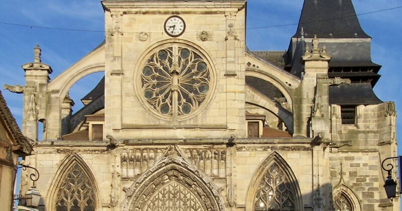 Église ancienne avec une façade en pierre, vitraux et horloge, sous un ciel bleu.