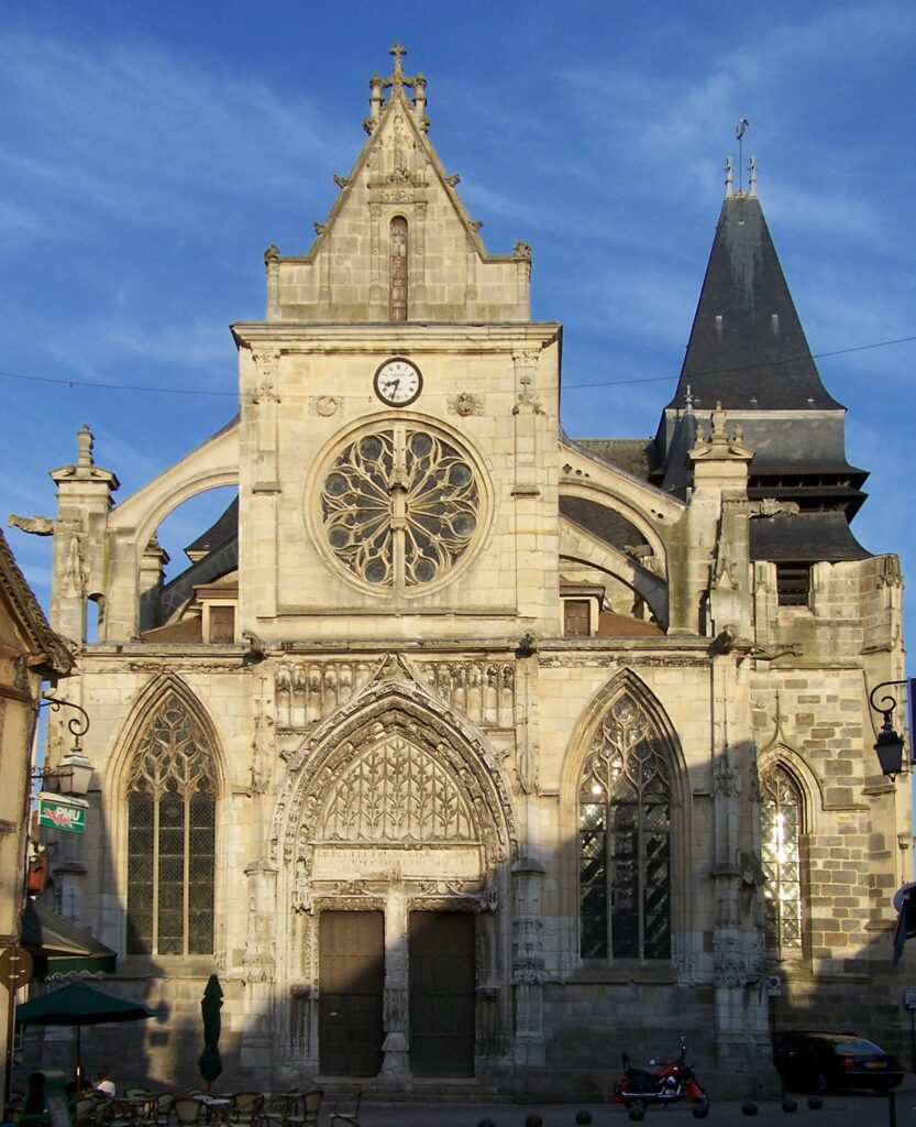 Église ancienne avec une façade en pierre, vitraux et horloge, sous un ciel bleu.
