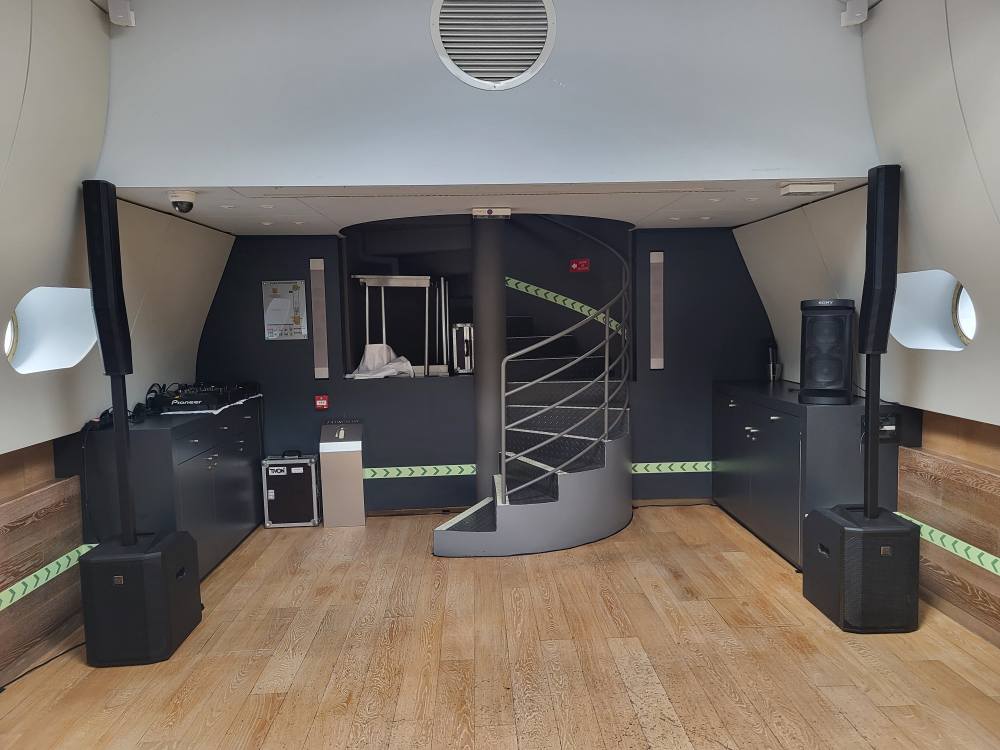 Intérieur d'une salle équipée pour le son et l'éclairage, avec tables, escaliers et matériel de location visible.