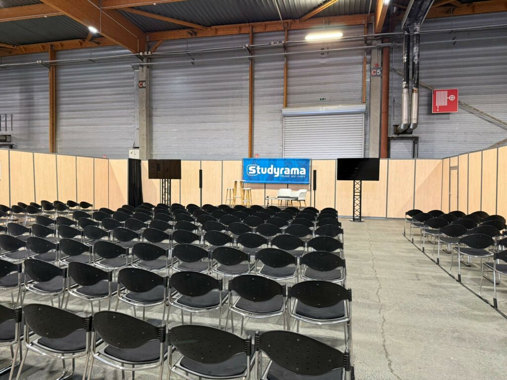 Salle de conférence avec rangées de chaises noires et espace pour un événement, avec un panneau "Studyrama" en arrière-plan.