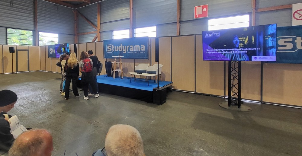 Stand de l'événement Studyrama avec une scène, des présentateurs et des participants en interaction dans un espace aménagé.