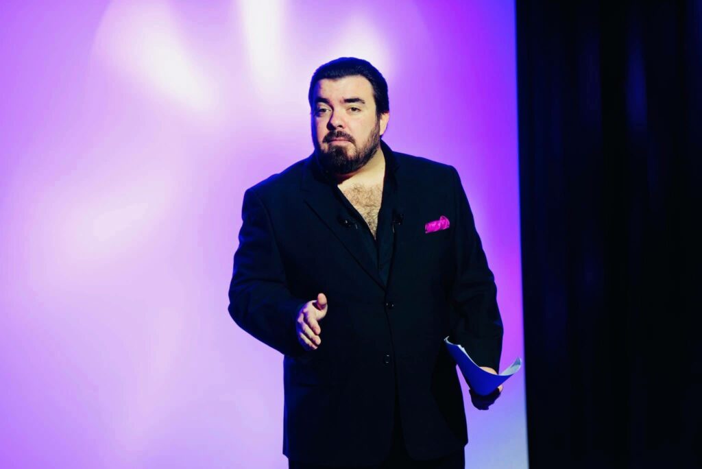 Homme en costume noir avec une chemise décolletée, tenant un papier, face à un fond violet.