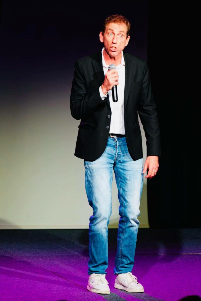 Un homme en costume noir avec une chemise blanche tient un micro sur scène, exprimant son énergie avec une gestuelle dynamique.