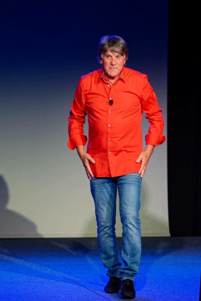Homme en chemise rouge sur scène, devant un fond sombre, se préparant pour une présentation.