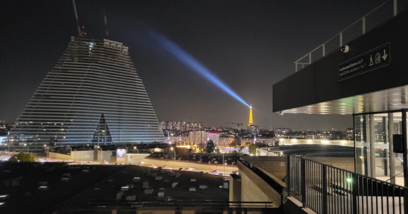 Vue nocturne de Paris avec la tour Eiffel illuminée, sur fond de bâtiment moderne.