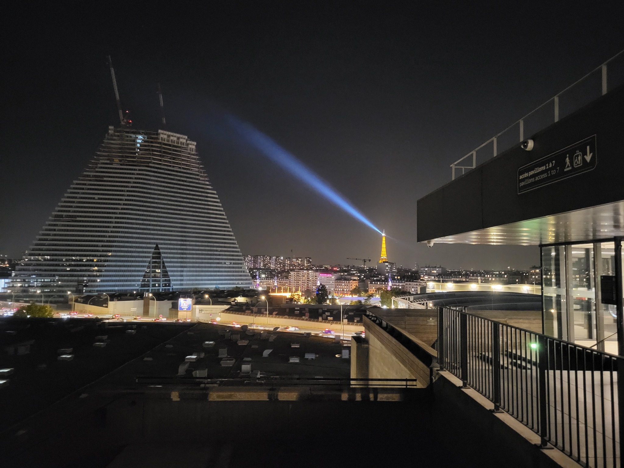 Vue nocturne de Paris avec la tour Eiffel illuminée, sur fond de bâtiment moderne.