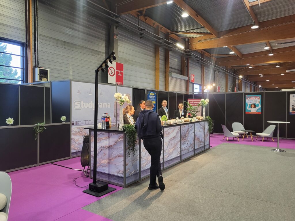 Stand d'exposition avec un comptoir, décor floral, et des personnes en conversation, dans un hall avec un sol violet.