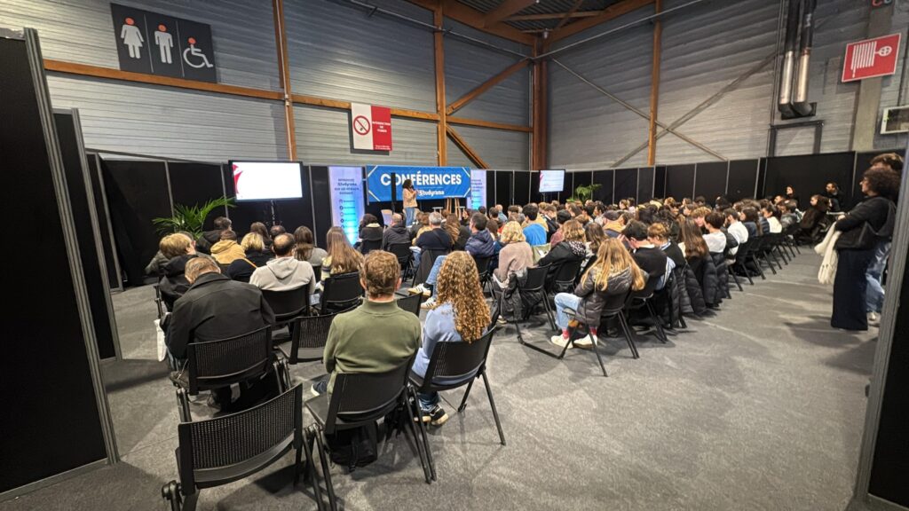 Public assistant à une conférence organisée par Symbiose Organisation, avec panneaux d'affichage et matériel audiovisuel en arrière-plan.