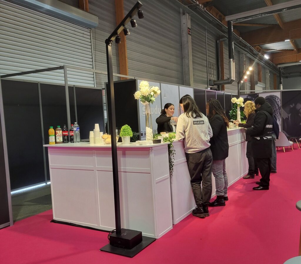 Un stand d'événement avec un bar animé, des boissons variées, des décorations florales et des personnes interagissant.