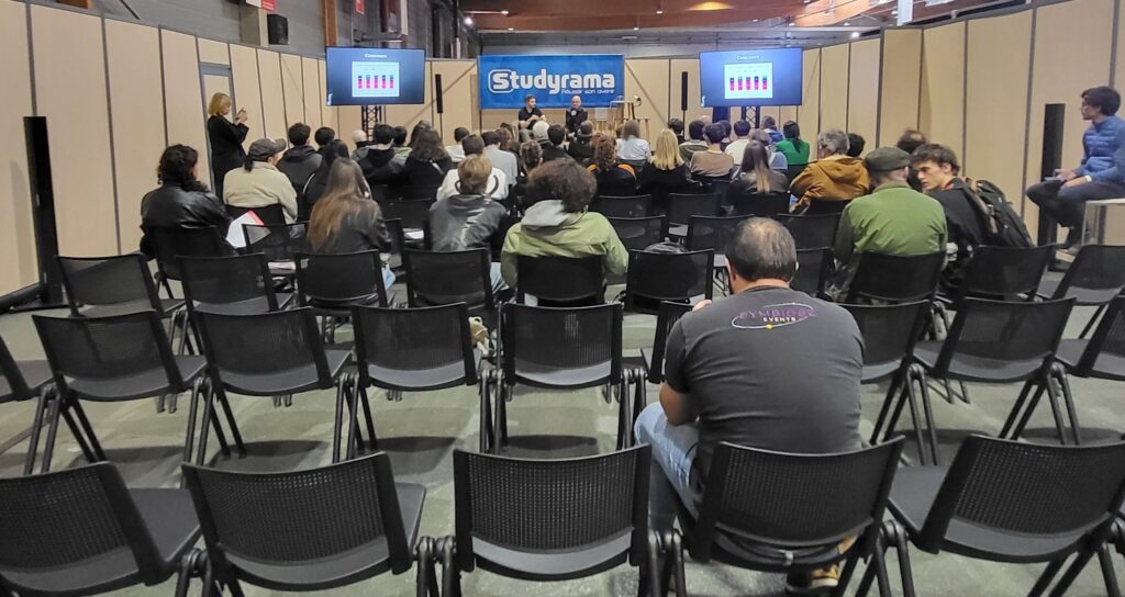 Audience attentive lors d'une présentation, avec des intervenants sur scène et des écrans à l'arrière.