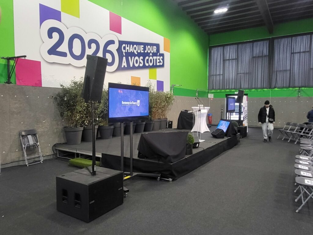 Installation de sono et éclairage pour un événement avec écran, scène et spectateurs, décorée de plantes.