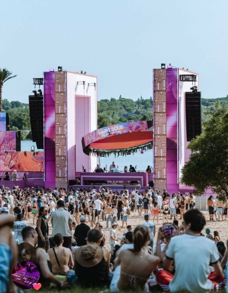 Scène colorée du festival Marvellous Island, avec une foule rassemblée sur la plage, entourée de matériel sonore et d'éclairage.