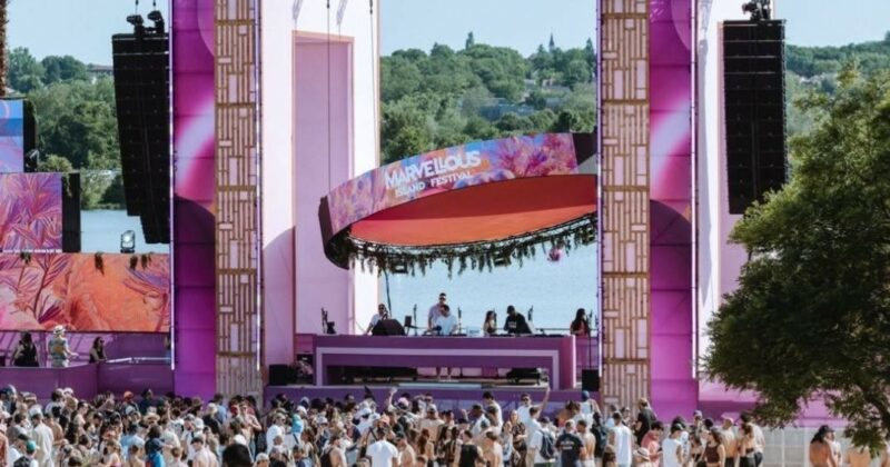 Scène colorée du festival Marvellous Island, avec une foule rassemblée sur la plage, entourée de matériel sonore et d'éclairage.