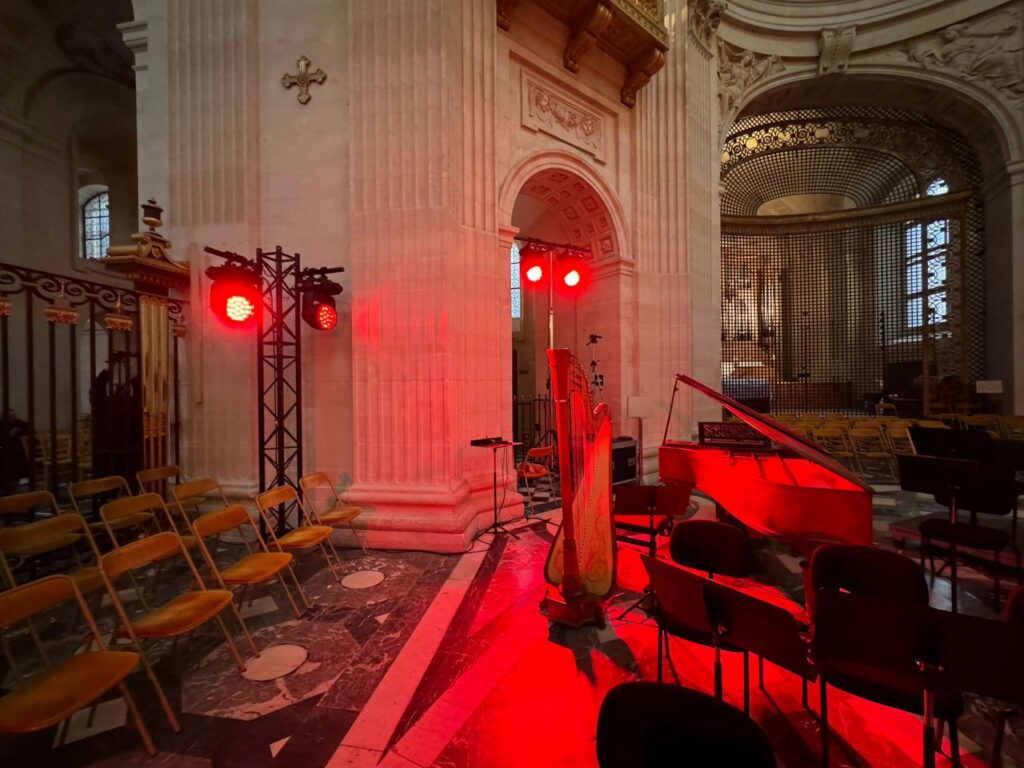 Matériel de son et éclairage installé, avec une ambiance rouge, dans un espace de concert. Harpe et piano en avant-plan.