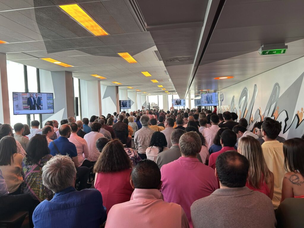 Une foule nombreuse assiste à une présentation dans une salle moderne, avec des écrans affichant des images.