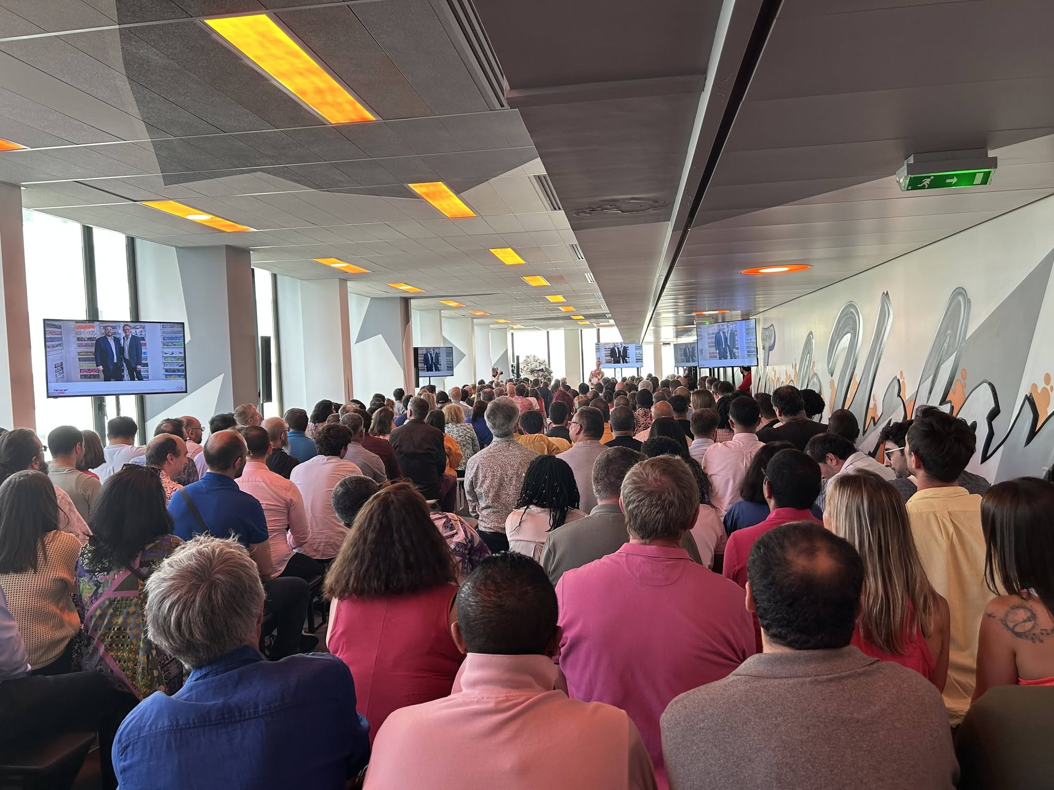 Une foule nombreuse assiste à une présentation dans une salle moderne, avec des écrans affichant des images.