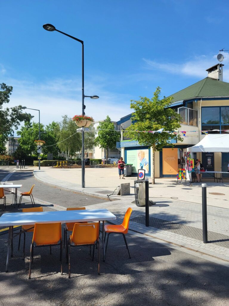 Espaces extérieurs équipés de tables et chaises orange, avec bâtiments et arbres en arrière-plan sous un ciel bleu.