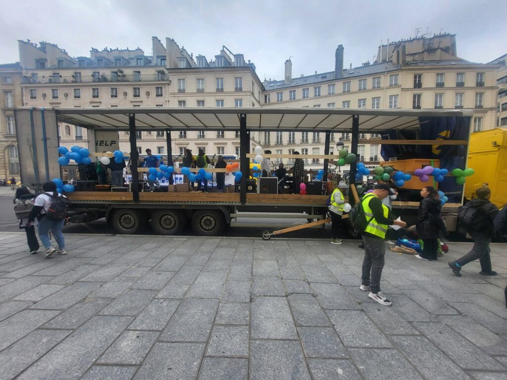 Camion de scène animé, décoré de ballons, avec des personnes en gilets jaunes, préparant un événement en plein air.