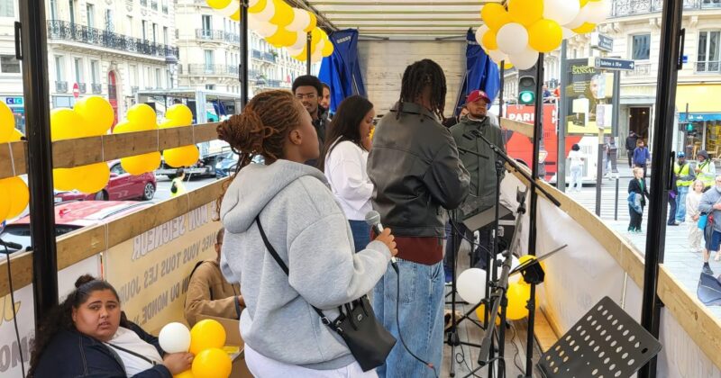 Des personnes chantent sur un camion, décoré de ballons jaunes, avec du matériel de sonorisation visible.