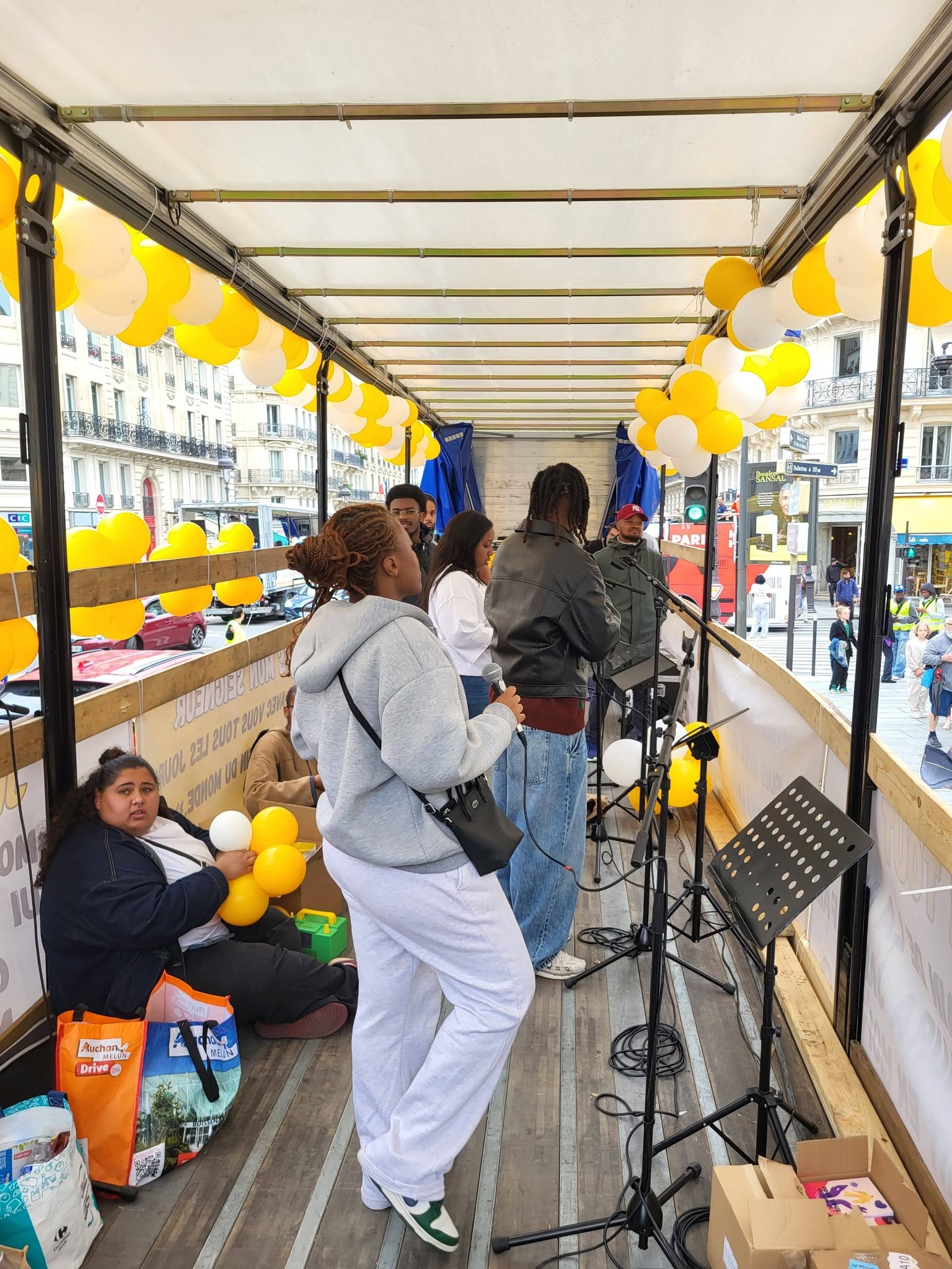 Des personnes chantent sur un camion, décoré de ballons jaunes, avec du matériel de sonorisation visible.