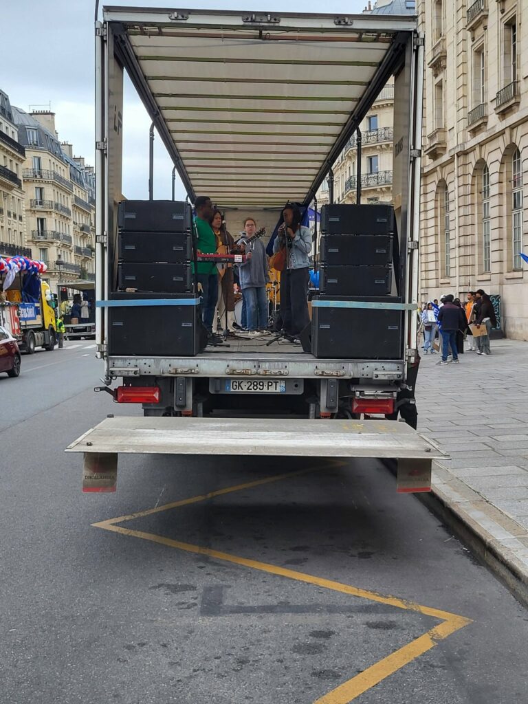 Groupe de musiciens sur une scène mobile, avec des équipement son et éclairage, lors d'un événement en centre-ville.