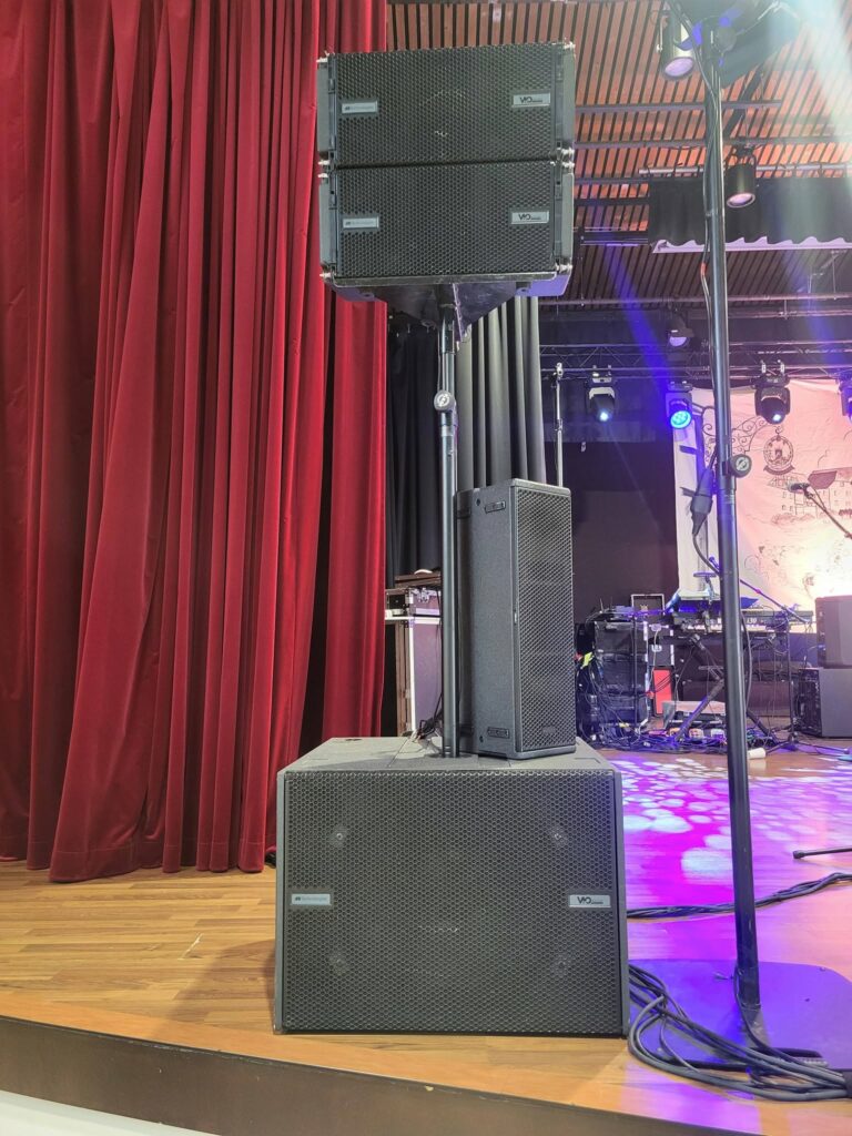 Système de sonorisation empilé sur scène, avec plusieurs enceintes devant un rideau rouge.