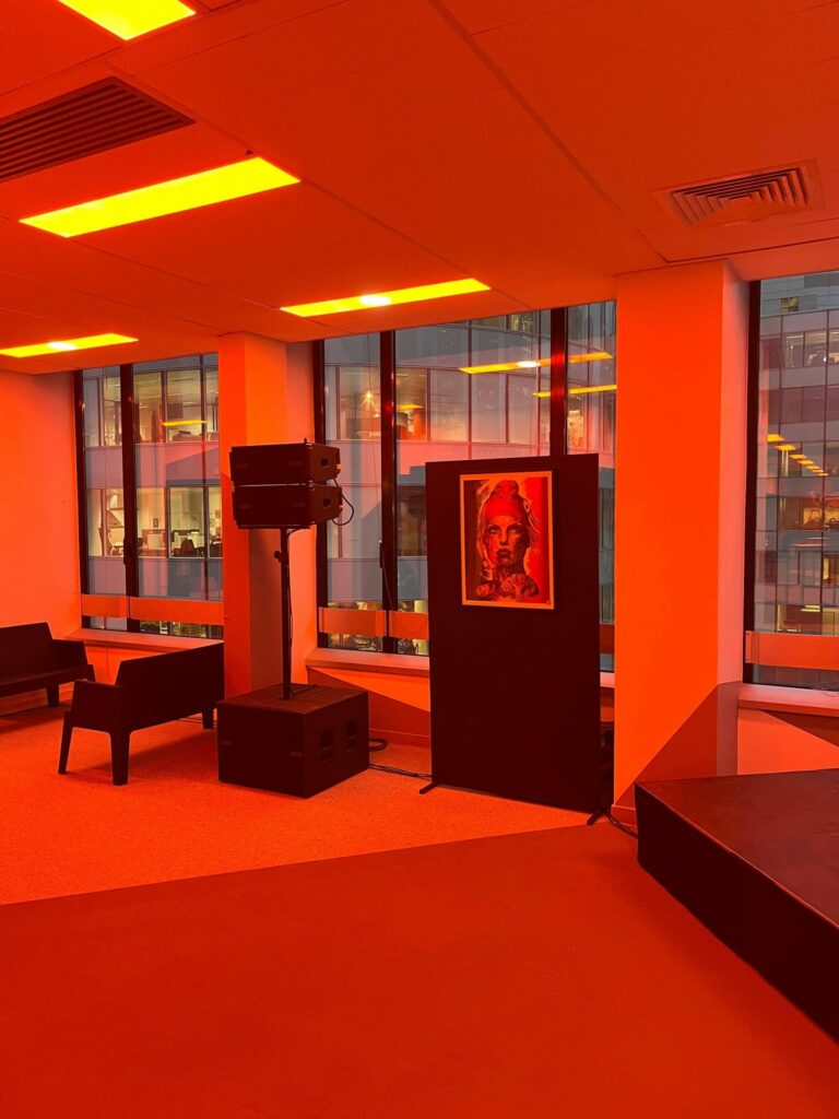Espace intérieur avec matériel de sonorisation et éclairage, éclairé en rouge, présentant une œuvre d'art sur un panneau.
