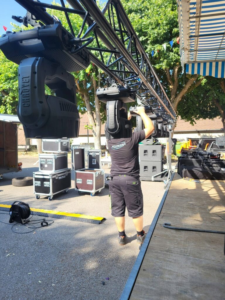 Technicien installant un système de sonorisation et d'éclairage sur une scène en plein air avec du matériel professionnel.