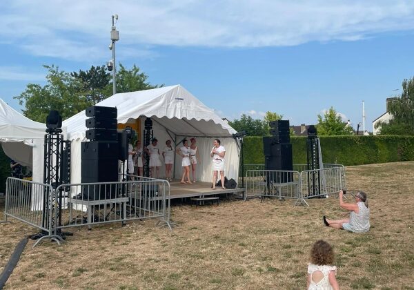Spectacle en plein air avec scène, matériel sono et éclairage installé, public assis sur l'herbe.