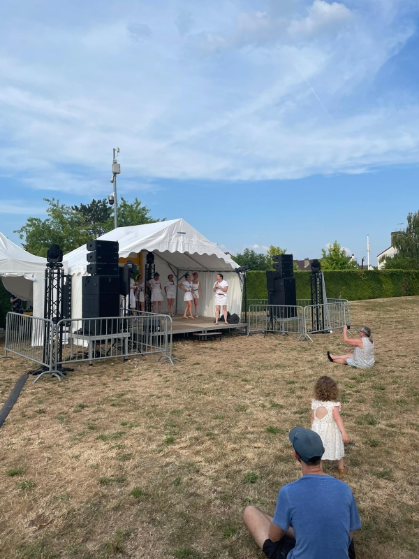 Spectacle en plein air avec scène, matériel sono et éclairage installé, public assis sur l'herbe.