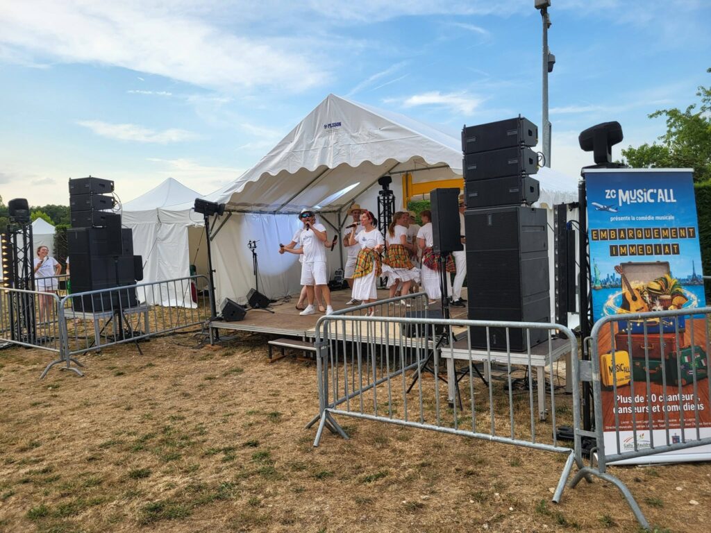 Scène de spectacle en plein air avec sonorisation et éclairage, représentant un groupe en performance. Tentes blanches en arrière-plan.