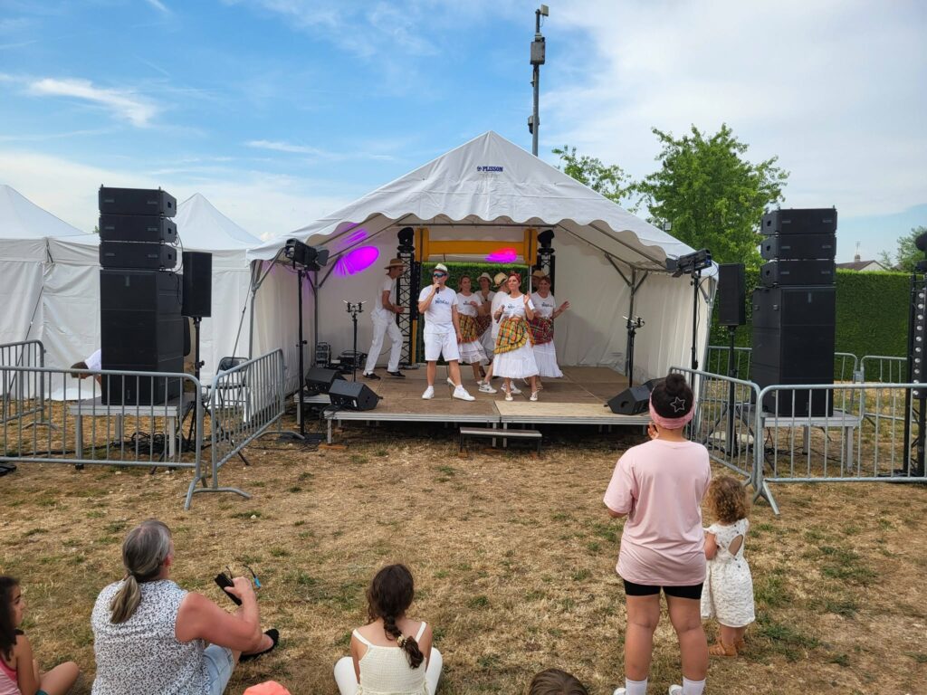 Spectacle en plein air avec un groupe sur scène et des spectateurs, entouré de matériel de sonorisation et d'éclairage.